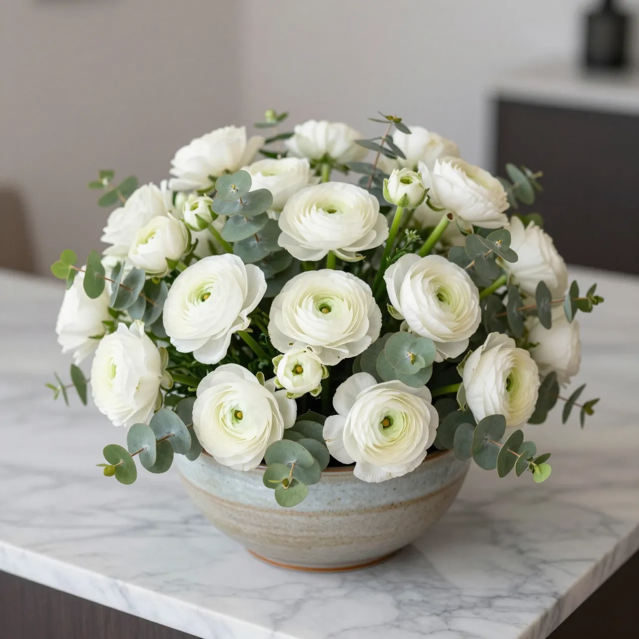 A lush low arrangement of white ranunculus and eucalyptus sits in a ceramic bowl on a marble countertop.