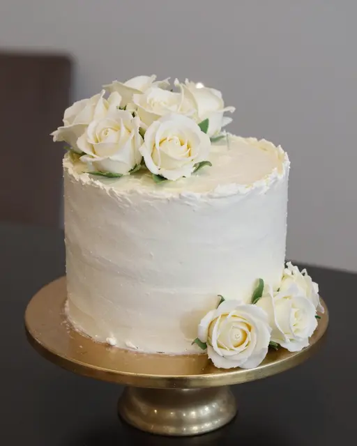 White buttercream cake decorated with sugar roses sits on a gold stand with a small lit candle