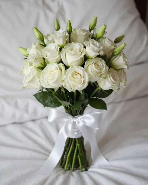 An anniversary bouquet of white roses and lisianthus tied with a satin ribbon rests on rumpled white bed linens.