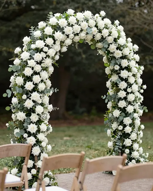Lush white rose and eucalyptus floral arch stands above wooden ceremony chairs in a garden.