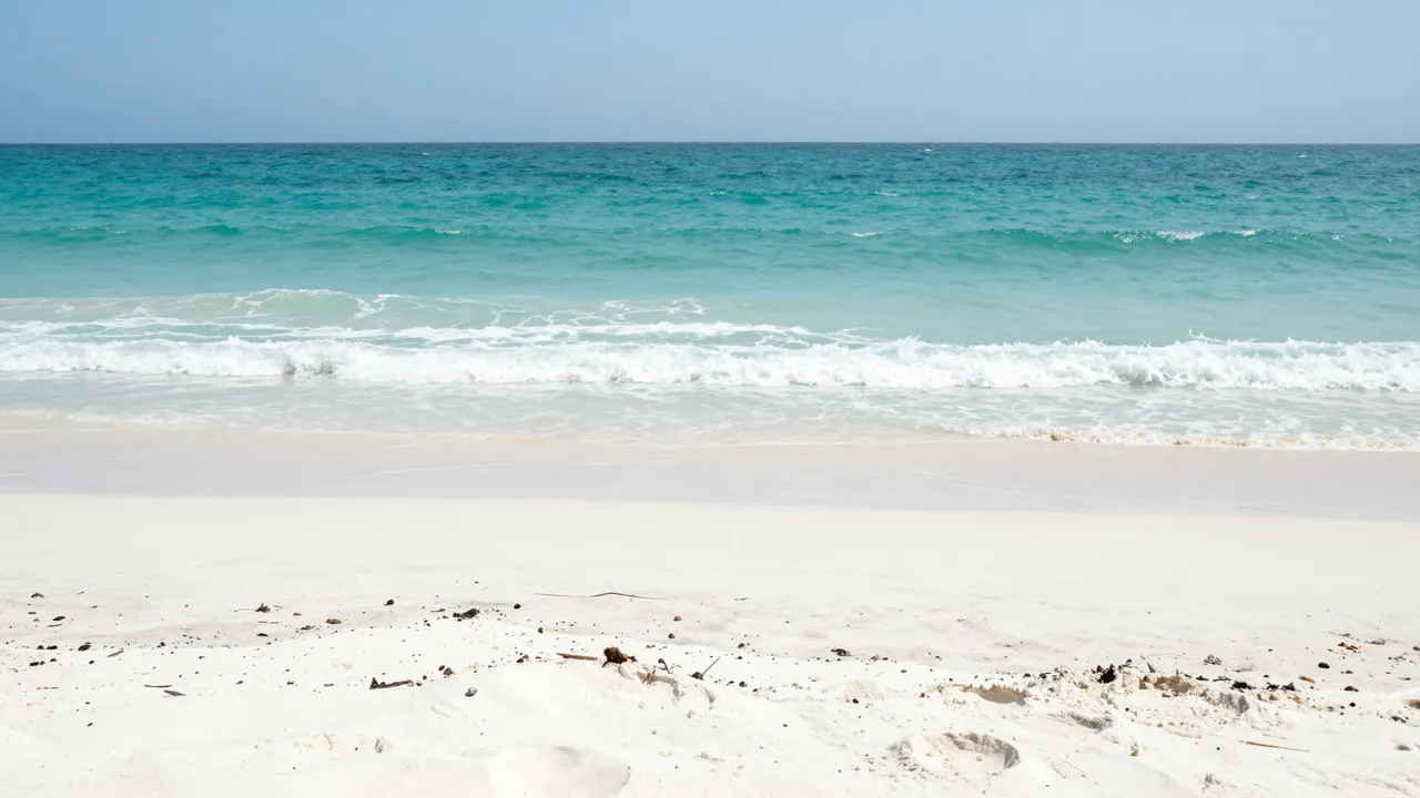 A quiet white sand beach with gentle turquoise ocean waves under a pale clear midday sky