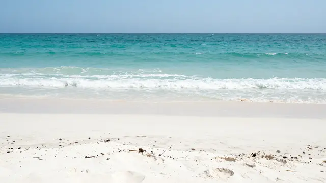 A quiet white sand beach with gentle turquoise ocean waves under a pale clear midday sky