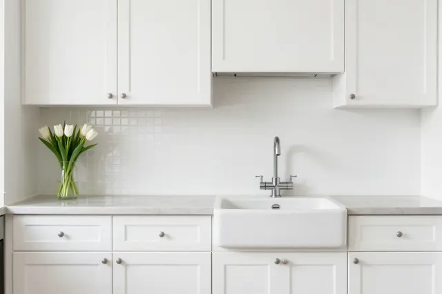 Bright minimalist white kitchen with shaker cabinets, farmhouse sink and a vase of white tulips on the countertop.