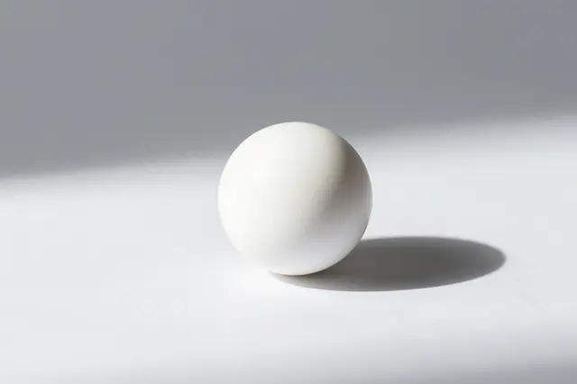 A smooth white sphere rests on a plain white surface casting a soft long shadow