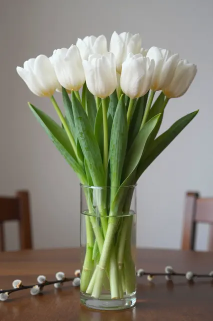 A clear glass vase of white tulips sits on a wooden table with pussy willow sprigs for Easter decor
