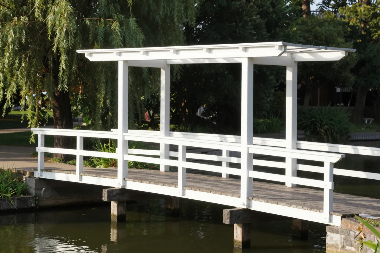 A covered white painted wooden footbridge spans a calm canal on a sunlit summer morning