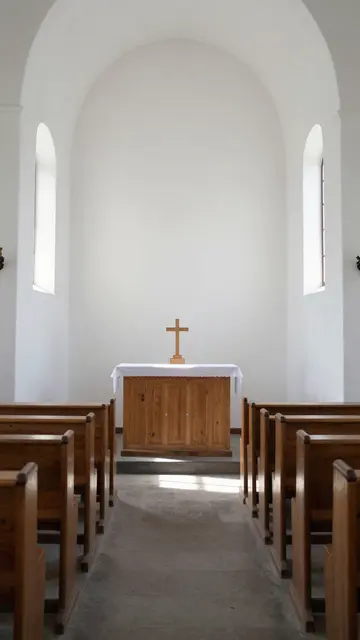 Sunlit aisle between wooden pews leads to a simple altar in a small white chapel