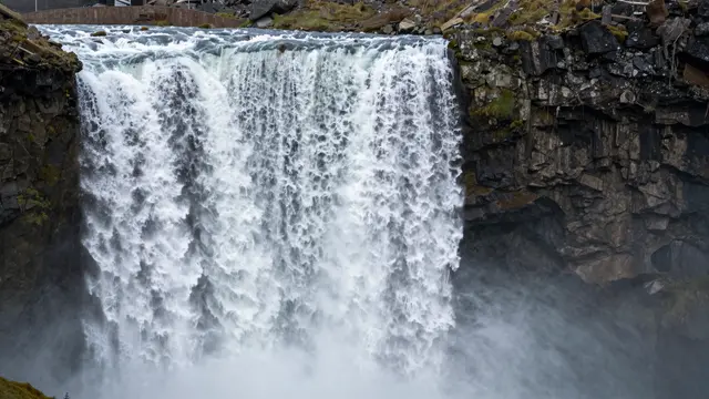 A broad sheet of rushing white water cascades over a dark mossy basalt cliff into mist below.