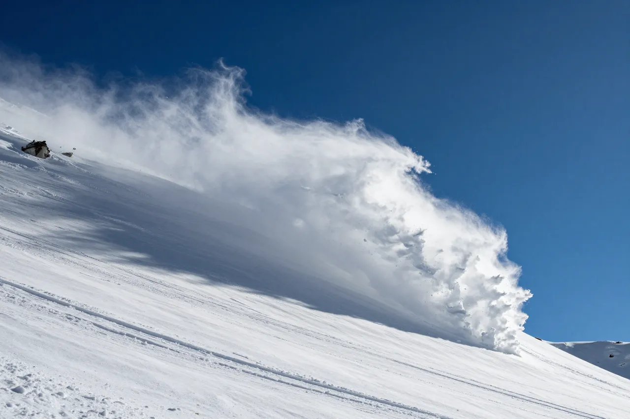 Wind carries plumes of blowing snow across a steep snowy mountain slope under a deep clear blue sky