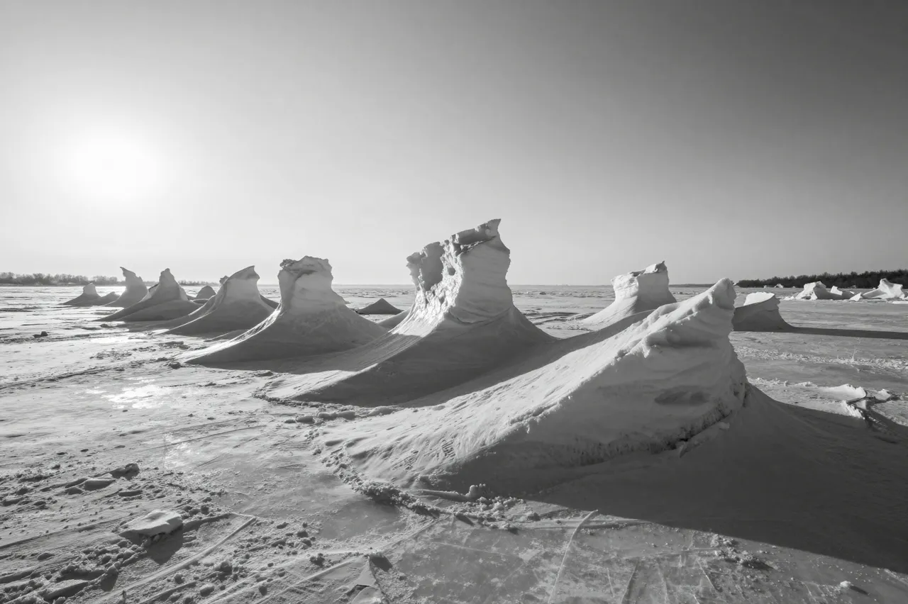 Black and white view of jagged wind-sculpted ice formations on a frozen lakeshore under a low bright sun