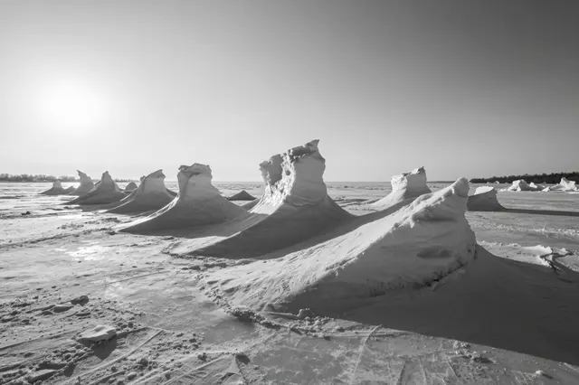 Black and white view of jagged wind-sculpted ice formations on a frozen lakeshore under a low bright sun