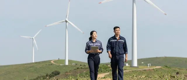 Two industrial technicians walk across grassy hills toward operating wind turbines