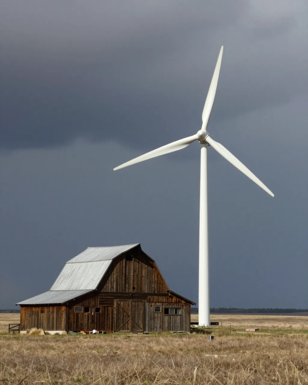 A white wind turbine stands next to an old wooden barn on a grassy prairie under dark storm clouds