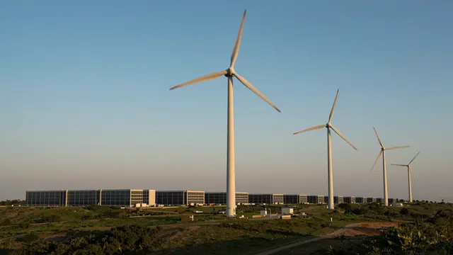 Row of wind turbines stands on a grassy ridge above a low modern data center at sunset