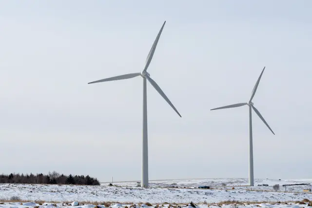 Two tall wind turbines stand in a snow-covered open plain beneath a pale overcast sky
