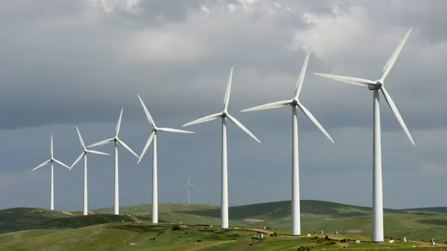A row of white wind turbines stands on green rolling hills under a dramatic cloudy sky