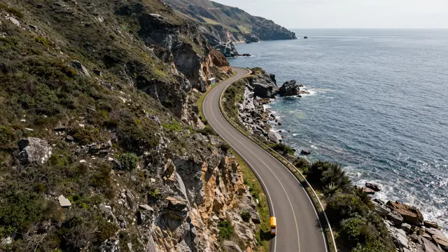Aerial view of a winding paved coastal road hugging rocky sea cliffs above a blue ocean