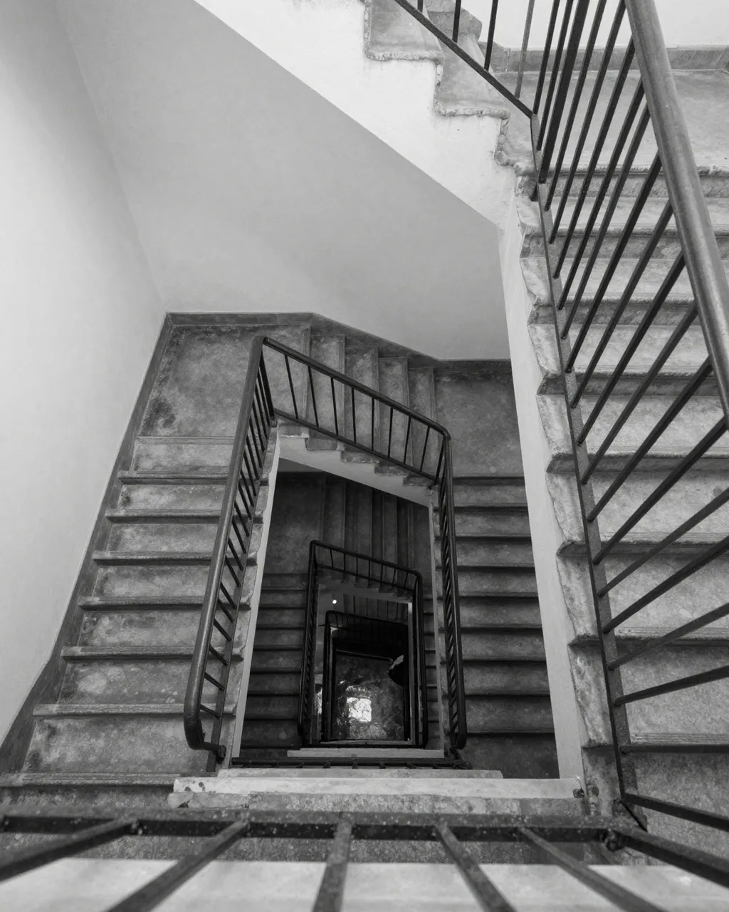 Black and white downward view of a stacked square concrete stairwell with iron railings in an old building