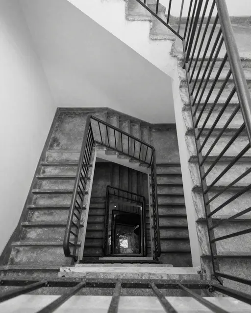 Black and white downward view of a stacked square concrete stairwell with iron railings in an old building