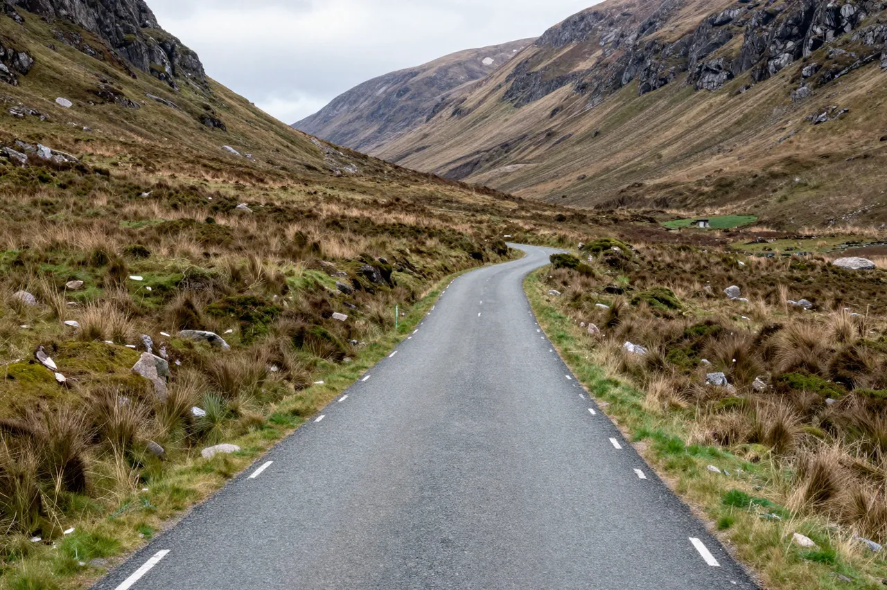 A single-track paved road winds through a rugged Scottish highland glen under overcast skies
