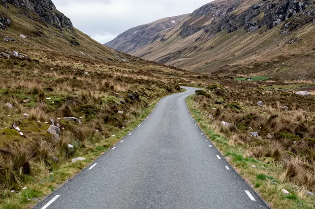 A single-track paved road winds through a rugged Scottish highland glen under overcast skies