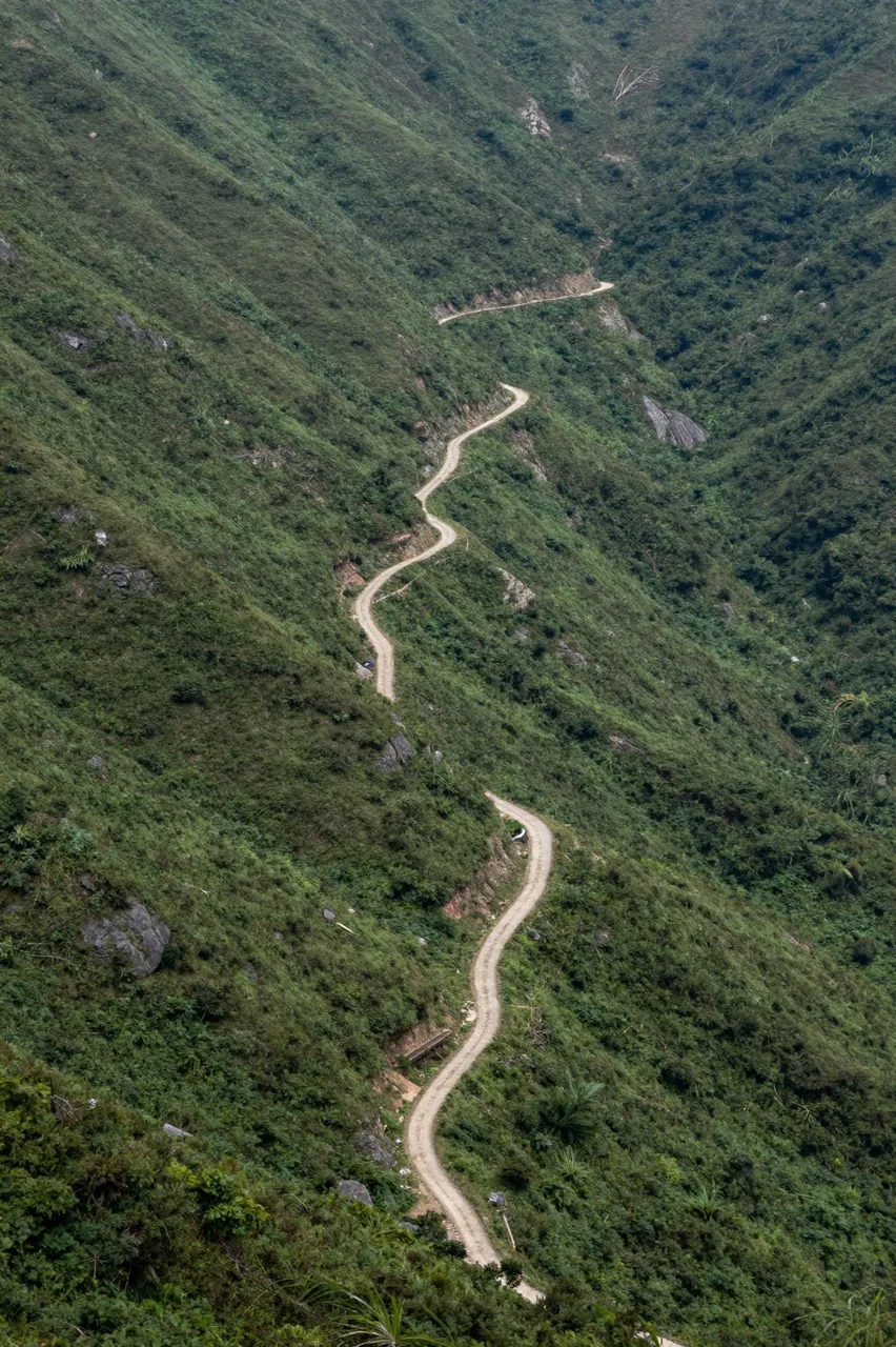 Aerial view of a narrow winding dirt road zigzagging up a steep green mountain slope