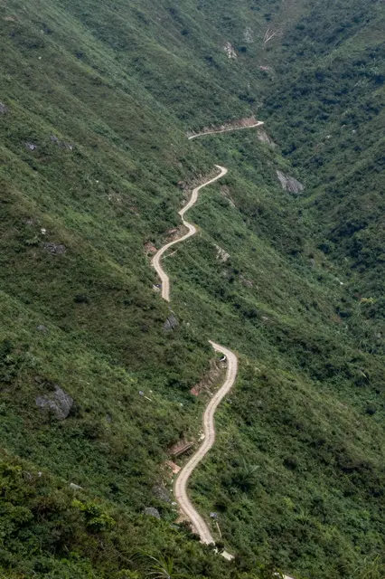 Aerial view of a narrow winding dirt road zigzagging up a steep green mountain slope