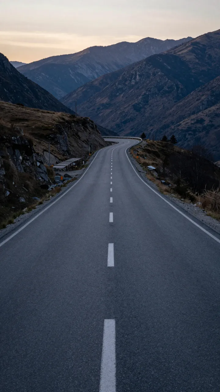 An empty paved road winds through rugged mountain ranges under a soft dusk sky
