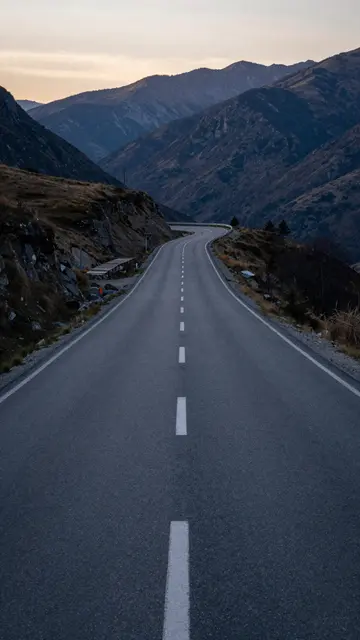 An empty paved road winds through rugged mountain ranges under a soft dusk sky