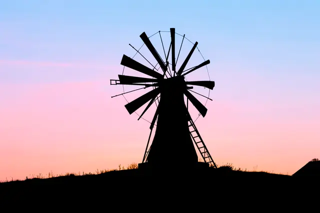 Black windmill silhouette stands on a low hill against a soft pastel pink and blue dawn sky