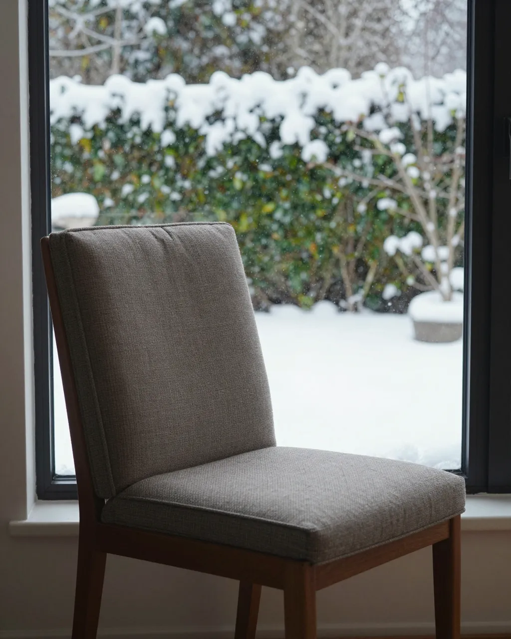 A upholstered wooden chair sits on an interior windowsill looking out at a snow-covered garden
