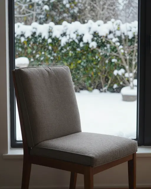 A upholstered wooden chair sits on an interior windowsill looking out at a snow-covered garden
