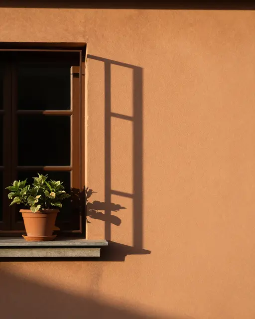 Warm evening sun casts window frame and potted plant shadows across a terracotta plaster wall with empty copy space to the