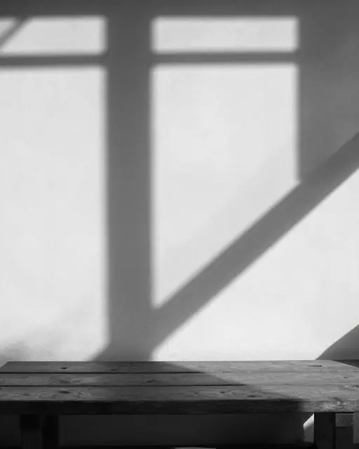 Black and white view of window frame shadows stretching across a white wall above a wooden bench in afternoon light.