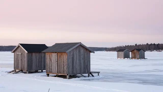 Weathered wooden fishing huts sit on a snow-covered frozen lake under a pale pink overcast sky