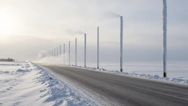 Paved high plateau road stretches through snow with tall snow-covered marker poles and blowing wind under a low pale sun