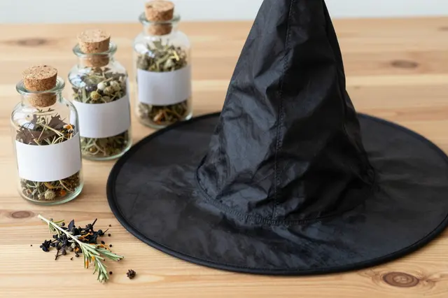 Black fabric witch hat sits beside unlabeled corked herb jars on a light wooden table