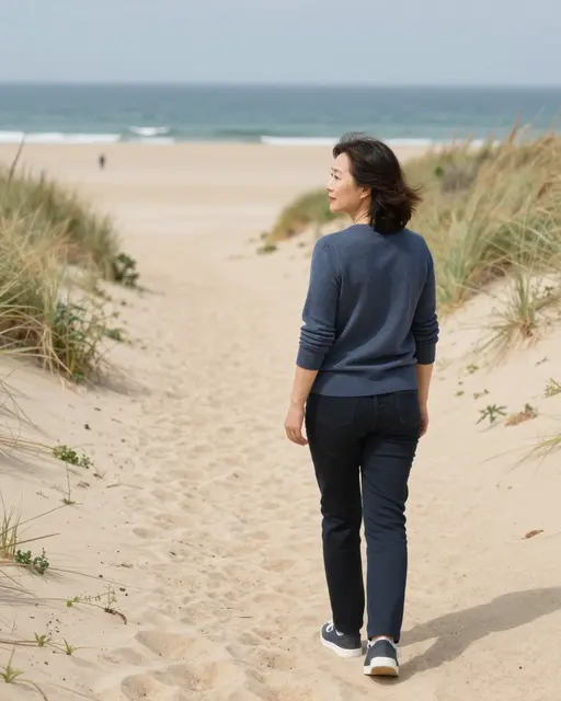 East Asian woman in her forties stands on a sandy dune path looking toward the beach and ocean