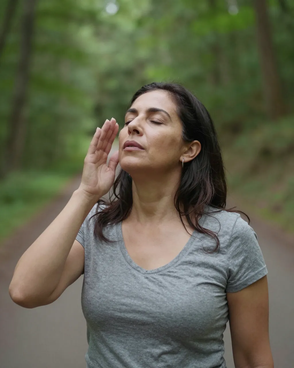 Middle aged Latina woman stands on a forest path with eyes closed, holding one hand near her face breathing slowly.