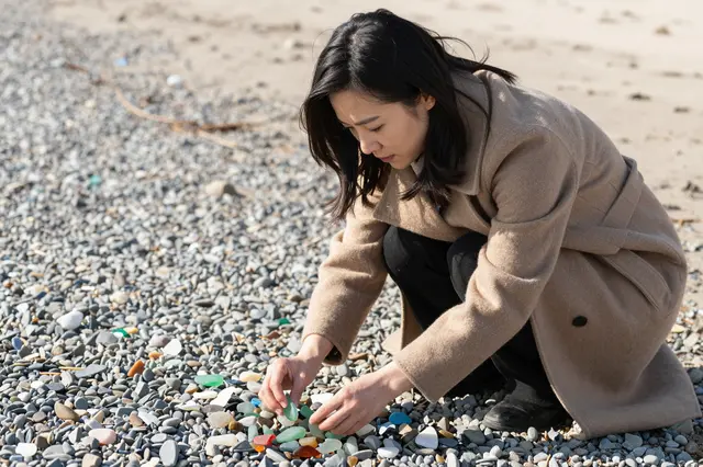 A Southeast Asian woman in a camel wool coat crouches on a pebbly shore sorting colorful sea glass in bright winter sunlight.