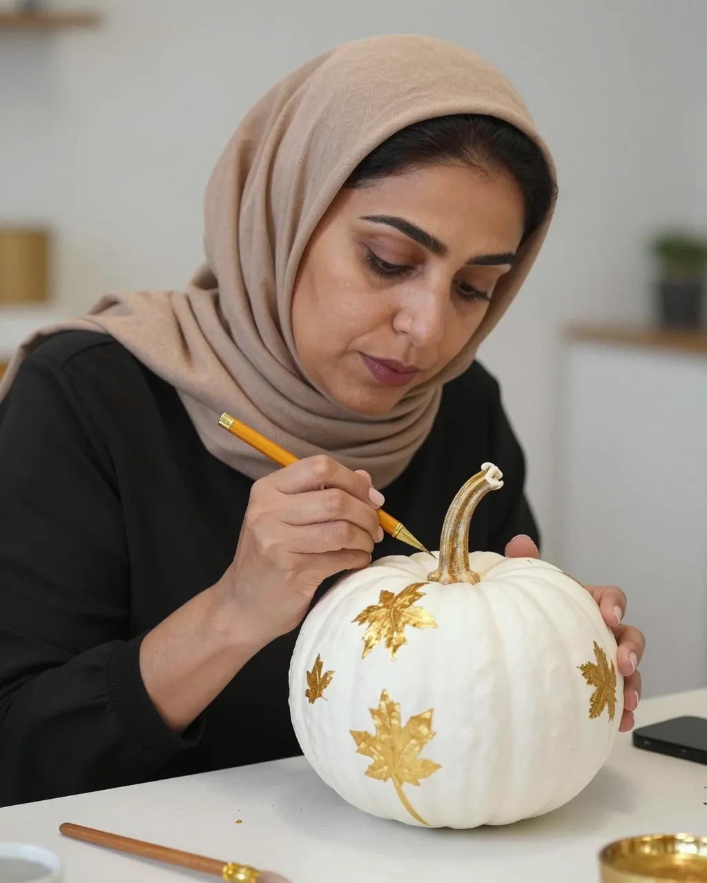 Middle Eastern woman in a headscarf paints gold leaf details on a white pumpkin at a craft table.