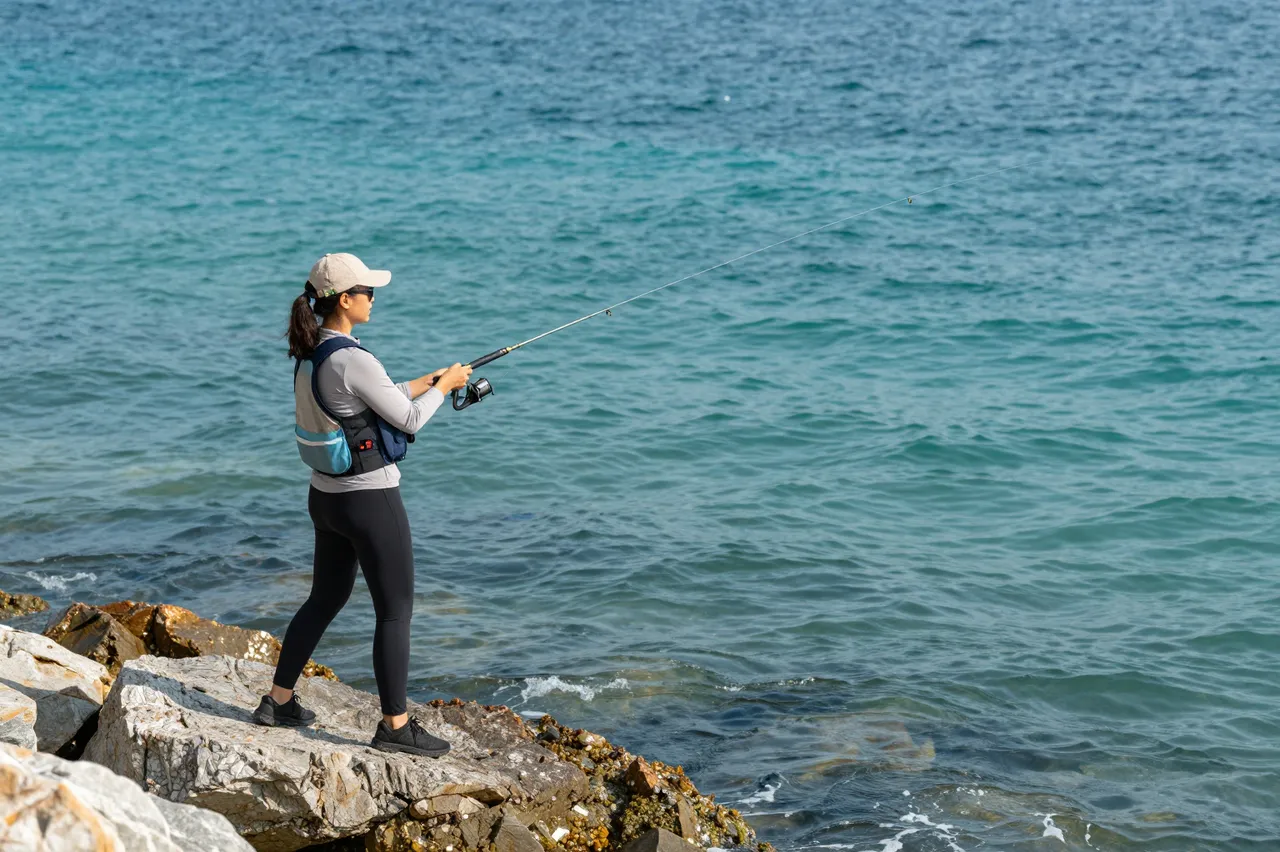 Side view of a woman standing on rocky shoreline casting a fishing rod into turquoise sea