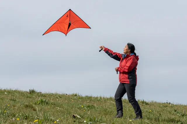 A middle-aged woman in a red jacket flies a bright red kite on a grassy hilltop under an overcast sky