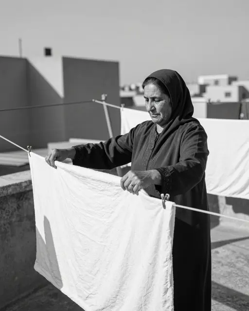 A mature Middle Eastern woman folds white fabric on a sunny urban rooftop between clotheslines