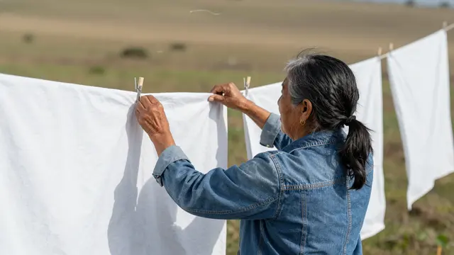 An older woman pins white sheets to an outdoor clothesline in a grassy field