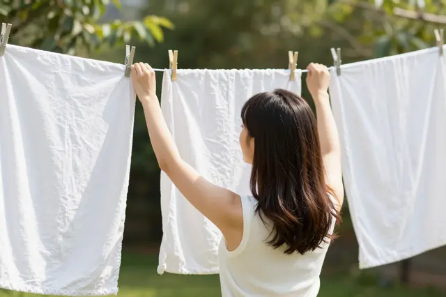 A woman viewed from behind hangs white linens on a clothesline in a sunny garden.