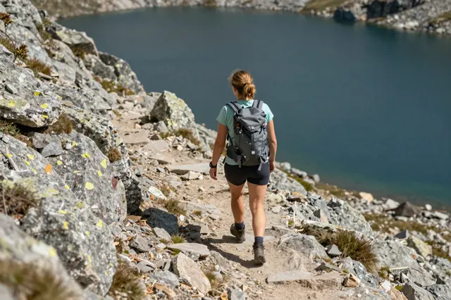 A woman viewed from behind hikes along a rocky mountain trail beside a deep blue alpine lake