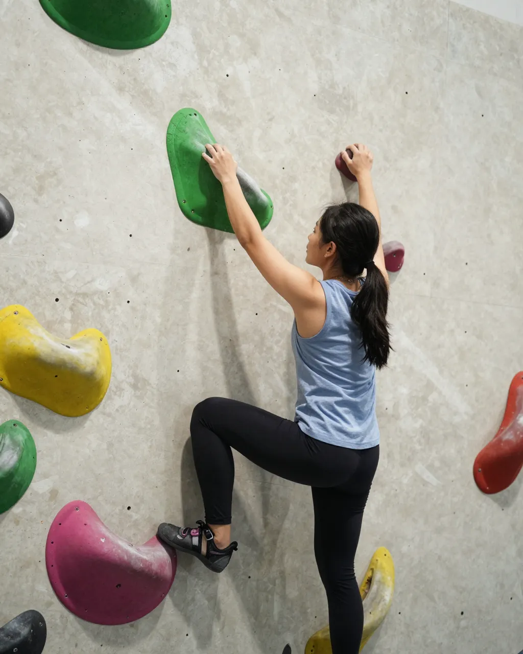 Back view of a young Southeast Asian woman gripping colorful holds on an indoor bouldering wall