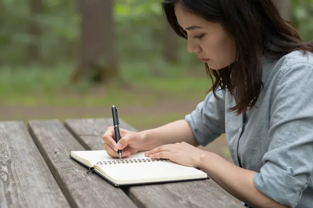 Side view of a woman writing in a blank spiral notebook at a wooden picnic table in a forest.