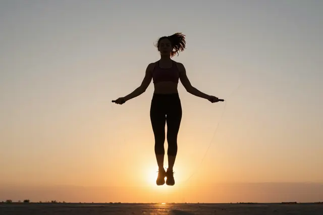 Silhouette of a woman mid-jump while holding a jump rope against a warm sunset sky
